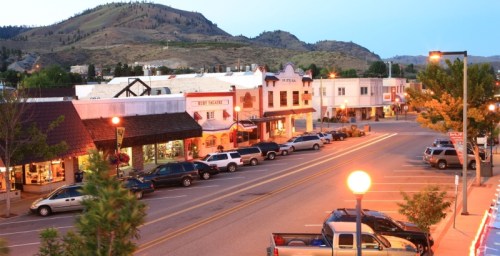 A quiet small town street at dusk, lined with shops and parked cars, with mountains in the background.