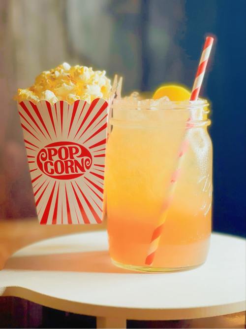 A striped popcorn box next to a mason jar filled with a colorful drink and a straw, set on a light surface.