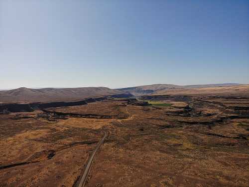 Aerial view of a vast, arid landscape with rolling hills and a winding road through a canyon. Clear blue sky above.