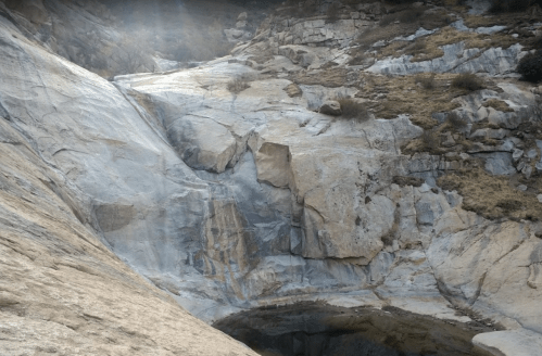 A rocky landscape featuring a waterfall cascading down smooth stone surfaces, surrounded by dry vegetation.