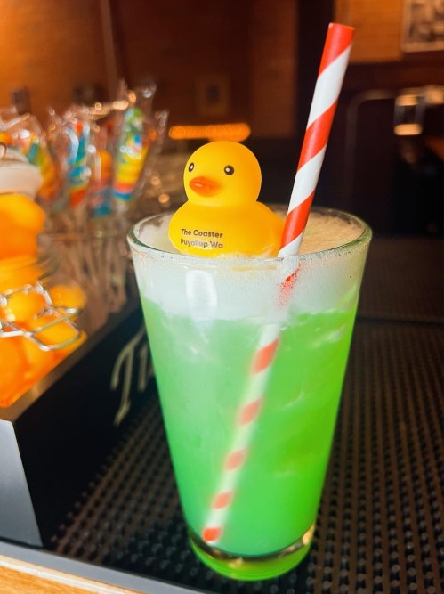 A green drink with a straw and a rubber duck decoration on top, set against a backdrop of colorful candy.