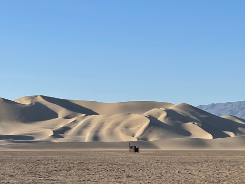 A small structure stands alone in a vast desert landscape with rolling sand dunes under a clear blue sky.