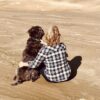A person in a plaid shirt sits on the beach, embracing a brown dog while looking out at the ocean.