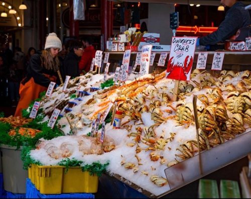A seafood market display featuring various crabs and shellfish on ice, with colorful signs and shoppers in the background.