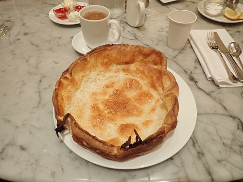 A large, golden-brown Yorkshire pudding on a white plate, with a cup of coffee and condiments in the background.
