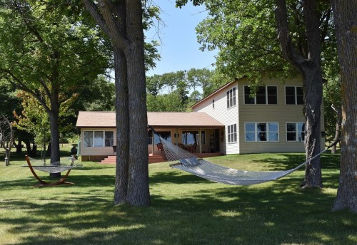 A spacious house surrounded by trees, featuring two hammocks in a grassy yard on a sunny day.