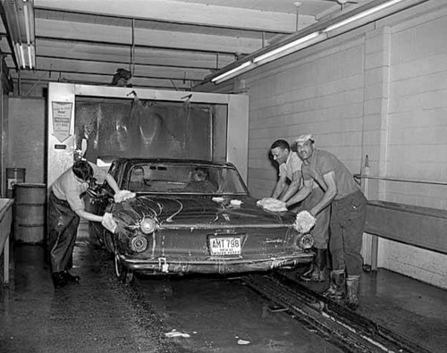 Three men wash a car in a vintage car wash, using sponges and water in a well-lit garage setting.