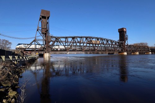 A train crosses a large, metal lift bridge over a river on a clear blue day.