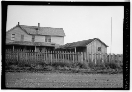 A black and white photo of a two-story house with a smaller adjacent building, surrounded by a wooden fence.