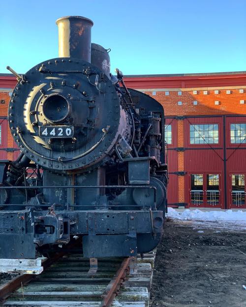 A vintage steam locomotive numbered 4420, parked on tracks in front of a red brick building.
