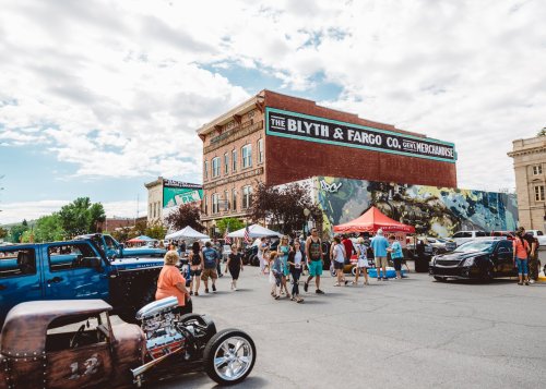 A bustling street scene with people, tents, and classic cars in front of a historic building with a mural.