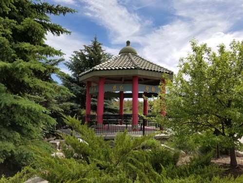 A gazebo with a tiled roof surrounded by lush greenery and trees under a partly cloudy sky.