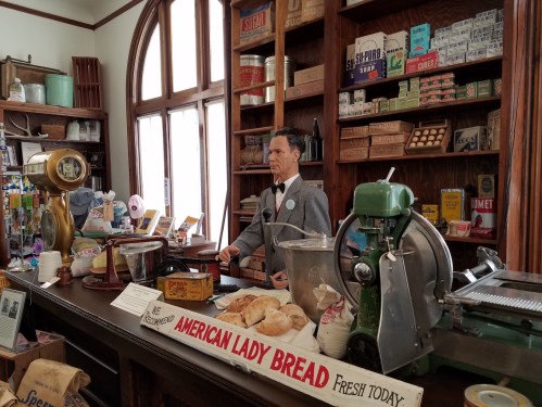 A vintage store display featuring a mannequin at a counter with old-fashioned goods and a sign for American Lady Bread.