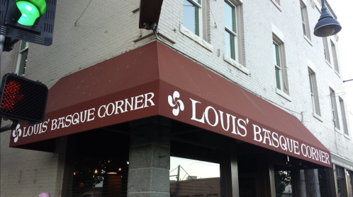 Sign for "Louis' Basque Corner" on a building with a red awning and a traffic light in the foreground.