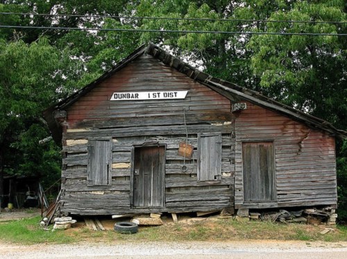 A weathered wooden building with a sign reading "Dunbar 1st Dist" surrounded by greenery and gravel.
