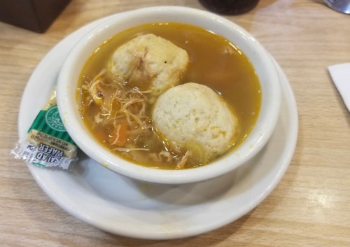 A bowl of chicken soup with two dumplings, garnished with shredded chicken and vegetables, served on a wooden table.