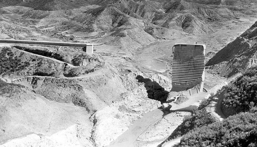 Black and white image of a large dam structure in a mountainous landscape, with a winding road nearby.