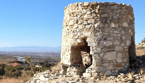 Stone ruins of a circular structure on a hillside, with a clear blue sky and distant mountains in the background.
