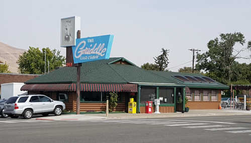 A roadside diner with a green roof and blue sign reading "The Griddle," surrounded by trees and parked cars.