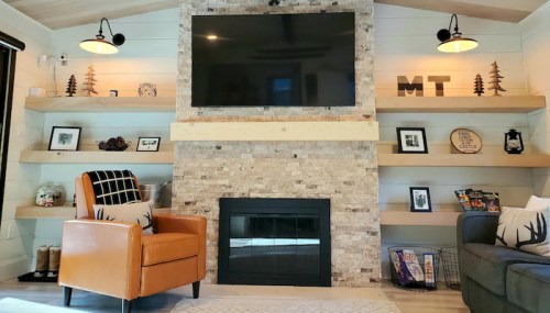 Cozy living room with a stone fireplace, TV, wooden shelves, and stylish decor, featuring an orange armchair and gray couch.
