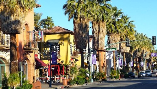 A sunny street lined with palm trees and colorful buildings, featuring outdoor dining and street signs.