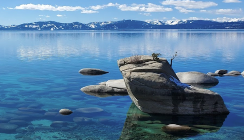 A serene lake scene featuring clear blue water, rocky formations, and distant snow-capped mountains under a bright sky.
