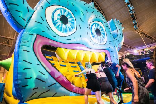 A colorful inflatable monster slide with people climbing up its side, set in a lively indoor environment.