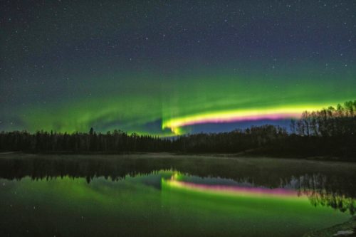 A vibrant aurora borealis lights up the night sky, reflecting in a calm lake surrounded by trees.