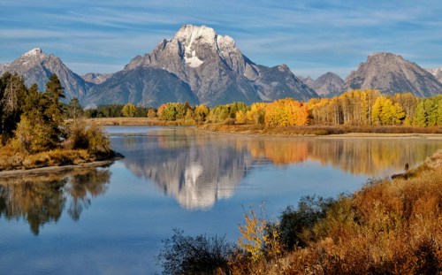 A serene landscape featuring a mountain range reflected in a calm river, surrounded by autumn foliage.