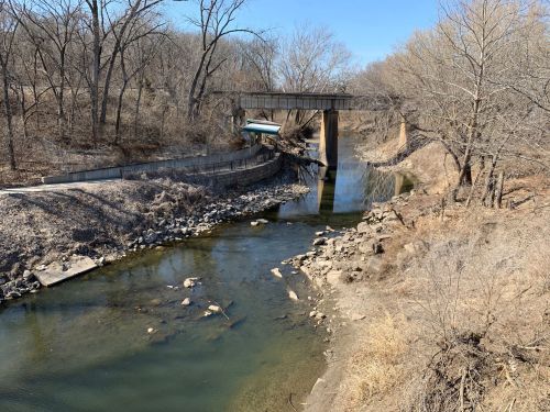 A serene river flows under a bridge, surrounded by bare trees and rocky banks on a clear day.