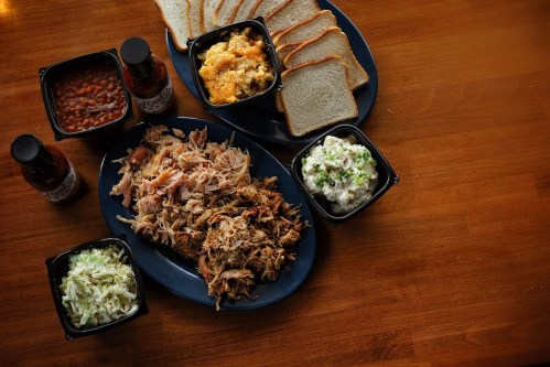 A platter of pulled pork, sides of coleslaw, potato salad, baked beans, and slices of bread on a wooden table.