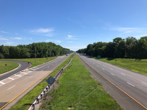 A wide view of a highway with green grass and trees on either side under a clear blue sky.