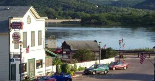 A scenic view of a riverside town with a hotel, parked cars, and flags, surrounded by lush green hills.