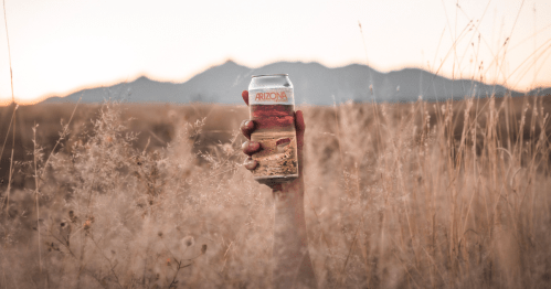 A hand holds a can of Arizona beer against a backdrop of mountains and tall grass at sunset.