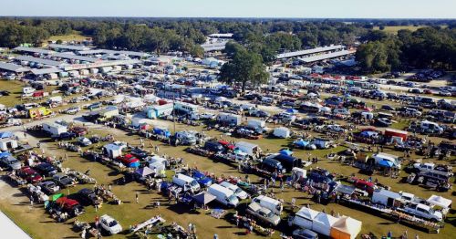 Aerial view of a bustling outdoor market with numerous tents, vehicles, and crowds under a clear blue sky.