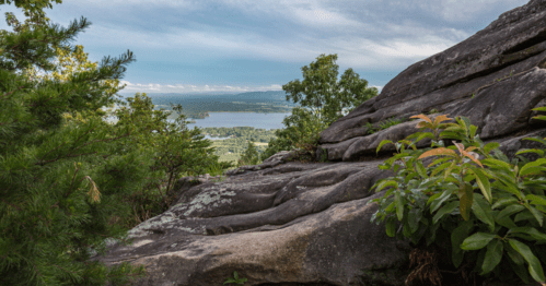 A rocky outcrop overlooks a lush landscape and a river, with trees framing the view under a cloudy sky.