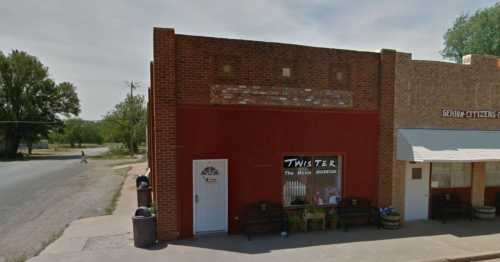 A brick building with a sign for the "Twister Movie Museum" and benches outside, located on a quiet street.