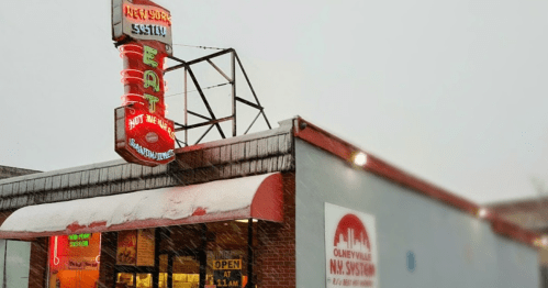 A retro diner sign glows in the snow, advertising "EAT" and "Hot Meat Sandwiches" on a chilly day.