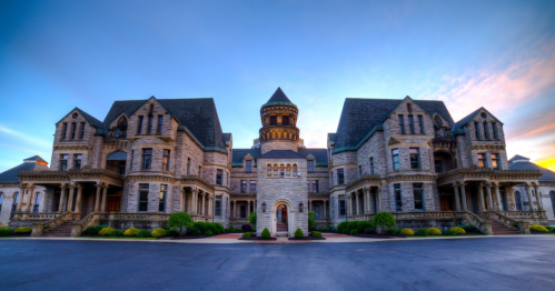 A grand, historic stone mansion with intricate architecture, featuring turrets and a symmetrical facade against a sunset sky.