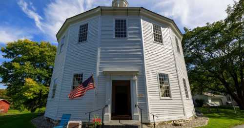 A round, white building with large windows, an American flag, and a welcoming entrance surrounded by greenery.