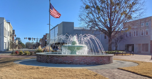 A fountain with water jets in a town square, with an American flag waving in the background and buildings nearby.