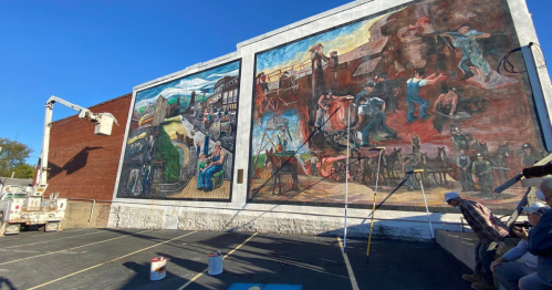 A large mural depicting historical scenes, with artists working on scaffolding beside it against a clear blue sky.