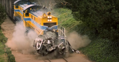 A train collides with a wrecked vehicle, creating a cloud of dust and debris on the tracks.