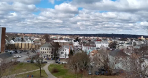 A panoramic view of a town with various buildings, trees, and a cloudy sky in the background.