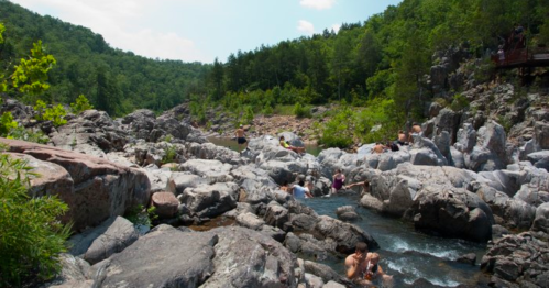 People relax and swim in a rocky river surrounded by lush green trees and hills under a clear blue sky.