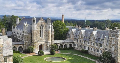 A scenic view of a stone building with gothic architecture, surrounded by green lawns and trees under a cloudy sky.