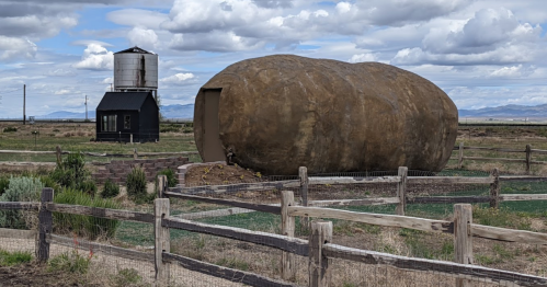 A large, potato-shaped structure sits in a field near a black building and a water tower under a cloudy sky.