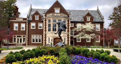 Historic brick mansion with a large statue in front, surrounded by colorful flowers and trees.