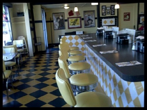 A retro diner interior featuring a checkered floor, yellow stools, and a long counter with vintage decor on the walls.