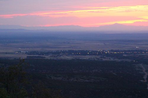 A scenic view of a valley at sunset, with colorful skies and distant mountains, illuminated by city lights below.
